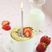 A Strawberry Shortcake in a Mug with a lit candle and sprinkles is beautifully garnished with fresh strawberries. Nearby, a carafe of milk and additional strawberries rest on the pink surface, creating a picture-perfect setting.