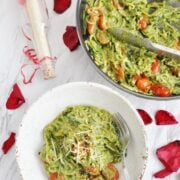 A bowl and pan of zucchini noodles with cherry tomatoes and a rich avocado pesto sauce, surrounded by red rose petals. Utensils rest in the pan while a fork stands ready in the bowl.