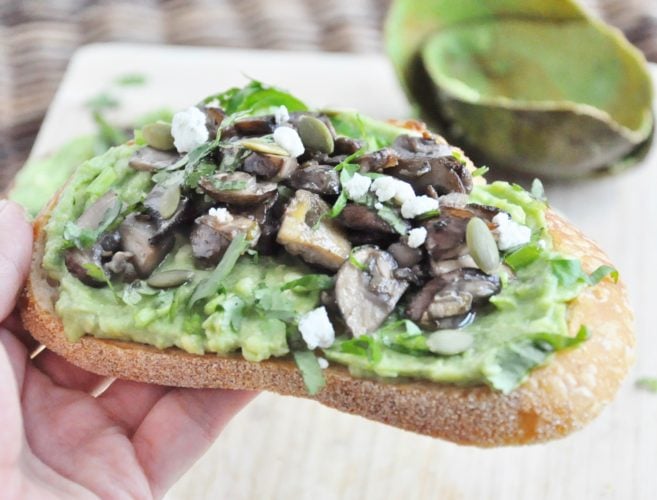 Avocado toast topped with sautéed mushrooms, a drizzle of truffle oil, crumbled cheese, pumpkin seeds, and herbs, all held delicately in hand.