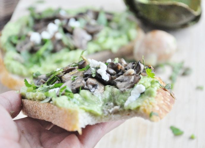 A hand presents a slice of avocado toast with sautéed mushrooms, crumbled cheese, herbs, and a hint of truffle oil. In the background, a softly blurred replica echoes this delicious composition.