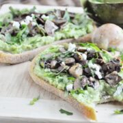 Two slices of bread topped with avocado and sautéed mushrooms, drizzled with truffle oil and sprinkled with feta, rest on a wooden board garnished with herbs. A round bread roll and part of an avocado complete the rustic scene.
