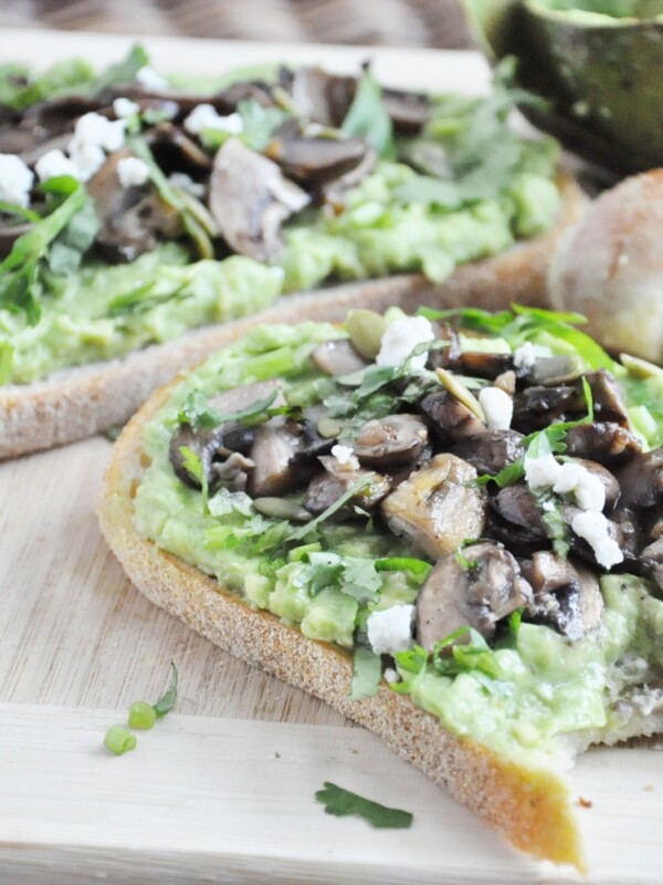 Two slices of bread topped with avocado and sautéed mushrooms, drizzled with truffle oil and sprinkled with feta, rest on a wooden board garnished with herbs. A round bread roll and part of an avocado complete the rustic scene.