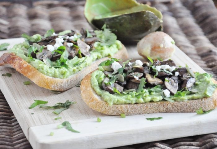 Two slices of bread topped with mashed avocado, sautéed mushrooms, and crumbled cheese, drizzled with luxurious truffle oil on a wooden board. In the background, avocado peel and pit complete this gourmet avocado toast setting.