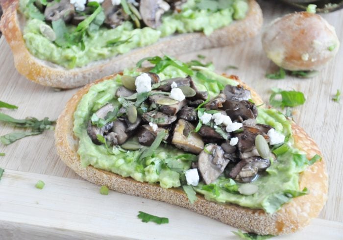 Avocado toast with sautéed mushrooms, a drizzle of truffle oil, crumbled cheese, pumpkin seeds, and chopped herbs served on a wooden board.