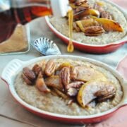 Two bowls of apple pie oatmeal with pecans are topped with sliced apples. Syrup is being poured over them from a bottle. A spoon and a jar of cinnamon are nearby, completing this cozy breakfast scene.