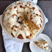 A Coconut Hummingbird Bundt Cake topped with white icing and pecan pieces graces a cake stand. Nearby, a slice rests on a plate, invitingly. A crisp white napkin peeks out beside the stand.