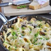 A pan of Mushroom and Spinach Pappardelle with grated cheese and herbs on top, featuring mushrooms and greens. In the background, a block of cheese, grater, and herbs rest on a cutting board.