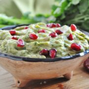A bowl of spicy chipotle guacamole topped with pomegranate seeds sits on a wooden surface, surrounded by fresh herbs and a chili pepper.