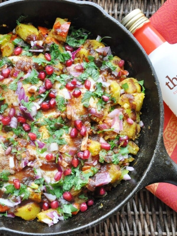 A skillet brimming with Chaat Masala Eggs and Potato Hash is adorned with vibrant pomegranate seeds, fresh herbs, and diced onions. Beside the skillet, a bottle labeled thrive and a red patterned cloth add charm to the scene.