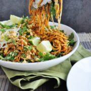 Tongs lifting a vibrant Tofu Pad Thai with sweet potato noodles, herbs, lime wedges, and vegetables in a white bowl on a green cloth.