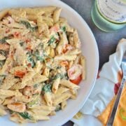 A bowl of creamy, spicy penne pasta with spinach and tomatoes, sprinkled with black pepper. A fork rests on a patterned napkin beside the dish. A bottle of white wine is partially visible.