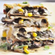 A stack of sliced vegetarian quesadillas with mushrooms, corn, onions, and cilantro is artfully arranged on a wooden surface. Delicately seasoned with truffle, these quesadillas bring an earthy richness to every bite.