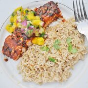 A plate with baked chipotle lime salmon topped with mango salsa next to a serving of brown rice garnished with cilantro. A fork rests on the white plates edge.