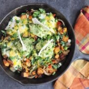 Skillet with eggs, melted cheese, and avocado on a flavorful fajita mix of veggies and potatoes. Topped with fresh herbs and served alongside a plaid cloth napkin.