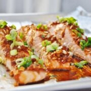 Three honey-glazed salmon fillets, infused with ginger and garlic, are topped with chopped green onions and sauce on a foil-lined tray.