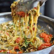 Caprese zucchini noodles tossed with cherry tomatoes and fresh herbs in a pan, elegantly lifted by metal tongs.
