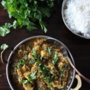 A bowl of Dhaniya Chicken Curry with herbs on top, a bowl of white rice, and fresh cilantro rest elegantly on a dark wooden table.