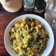 A bowl of Curry Roasted Butternut Squash Quinoa Salad sits on a wooden table, accompanied by a whole butternut squash and two spice jars. A napkin and utensils are neatly placed beside the vibrant plate.