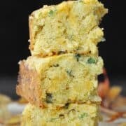 Three stacked pieces of jalapeño cheddar cornbread with green herb flecks rest gracefully against a dark background on a wooden surface, adorned with scattered autumn leaves.