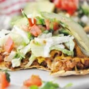 A Spicy Baked Turkey Tostada topped with ground turkey, melted cheese, shredded lettuce, diced tomatoes, chopped green onions, and avocado slices sits invitingly on a plate.