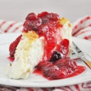 A slice of Cranberry Orange Cheesecake topped with a red cherry compote rests on a white plate, accompanied by a fork and checkered red and white cloth in the background.