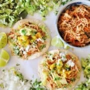 Two tacos topped with shredded chicken, diced onions, avocado, and cilantro evoke the rich flavors of Chipotle Chicken Tinga. Theyre surrounded by lettuce, lime wedges, and a bowl of chicken filling on a white surface. Perfect for those instant cravings!.