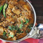 A bowl of Punjabi chicken curry garnished with cilantro and green chili, served in a metal dish on a red fabric background with a spoon nearby.