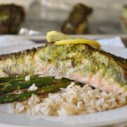 A plate of Hariyali Salmon features a baked salmon fillet with a green herb crust, served alongside white rice and asparagus, garnished with a fresh lemon slice.