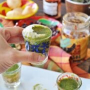 A hand holds a glass rimmed with spices and filled with a green sauce, reminiscent of the vibrant flavors of Mexican Pani Puri. In the background, a plate, bowls, and jars brimming with mixed condiments await discovery.