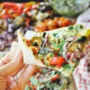 A hand holds a slice of vibrant veggie pesto flatbread pizza, topped generously with colorful vegetables. A red and white checkered cloth adds a touch of classic charm, with more food peeking through in the background.