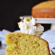 A slice of pistachio lemon cake with cardamom whipped cream, adorned with chopped pistachios, rests on a white plate. Another cake is in the background.