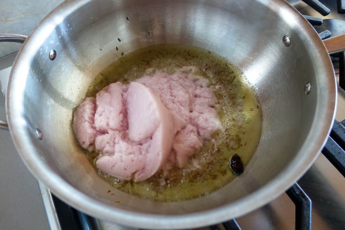 A stainless steel pan on a stovetop with pink blended onions frying in oil and whole spices.