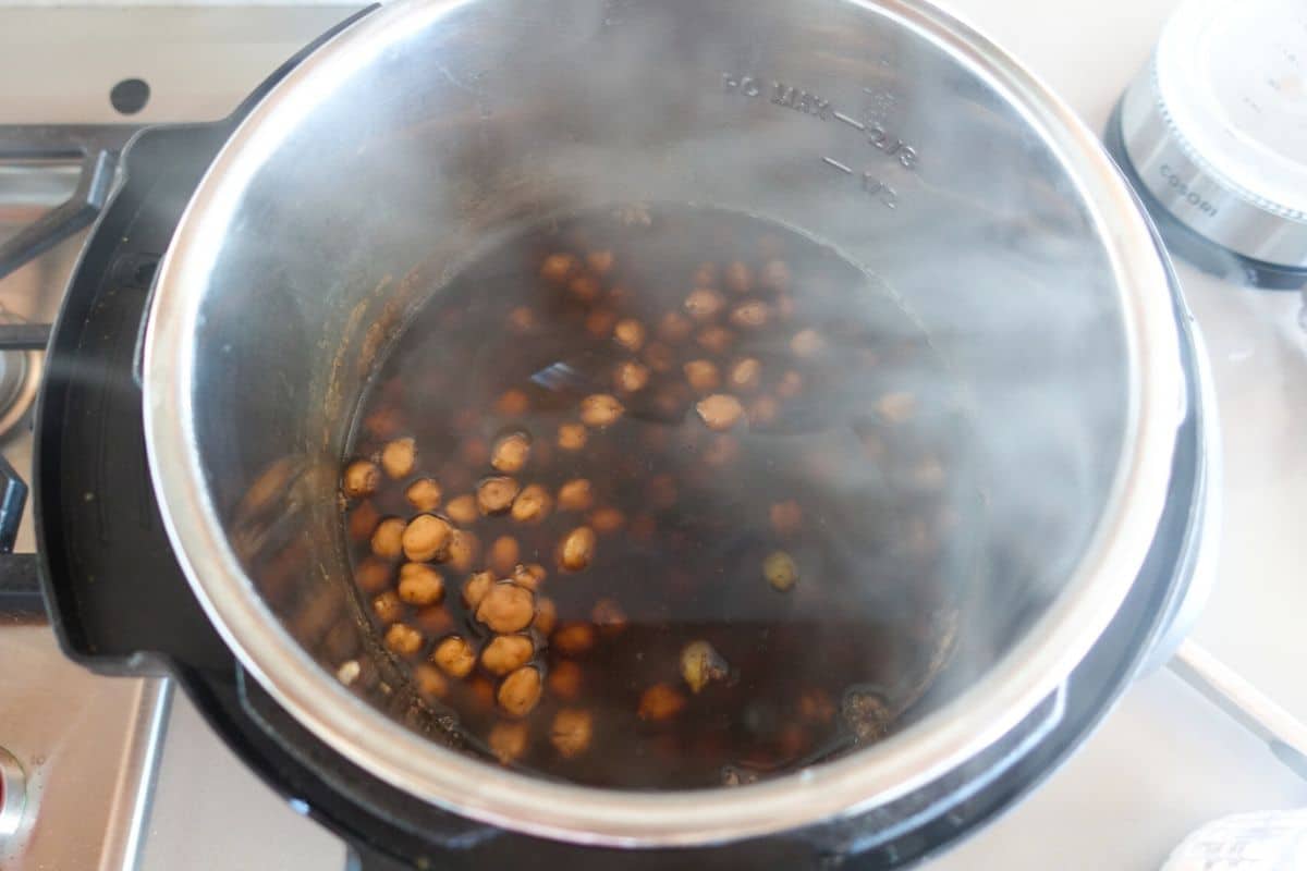 Cooked chickpeas simmering in dark liquid inside an open Instant Pot, with steam rising from the pot.