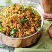 A bowl of Quinoa Broccoli Pulao mixed with fresh vegetables and garnished with cilantro, placed next to a small dish of pickle and a copper vessel.