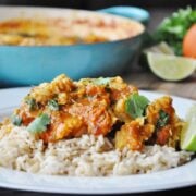 A plate of rice topped with Spicy Fish Masala curry garnished with cilantro, served with a lime wedge, with a tomato, lime, and a pot in the background.