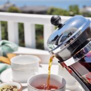 Coffee is poured from a French Press into a white cup on a table set with cups, snacks, and the aroma of Cardamom Fennel Chai, all against a scenic outdoor background.