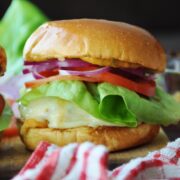 A Spicy Mexican Turkey Burger with lettuce, tomato, red onion, and melted cheese on a bun sits on a wooden surface beside a red and white striped cloth.