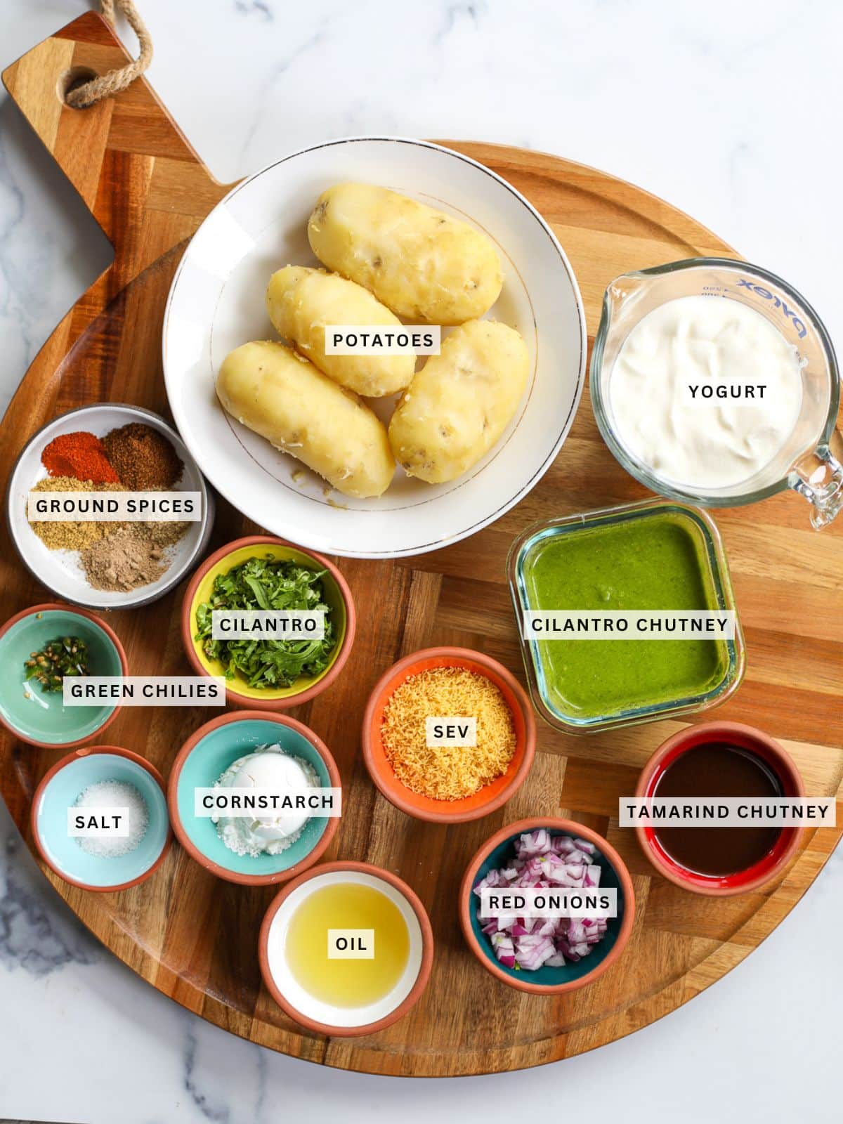 A round wooden board holds bowls of labeled ingredients for a delicious aloo tikki chaat, including potatoes, yogurt, ground spices, chutneys, sev, oil, salt, cornstarch, cilantro, green chilies, and red onions.