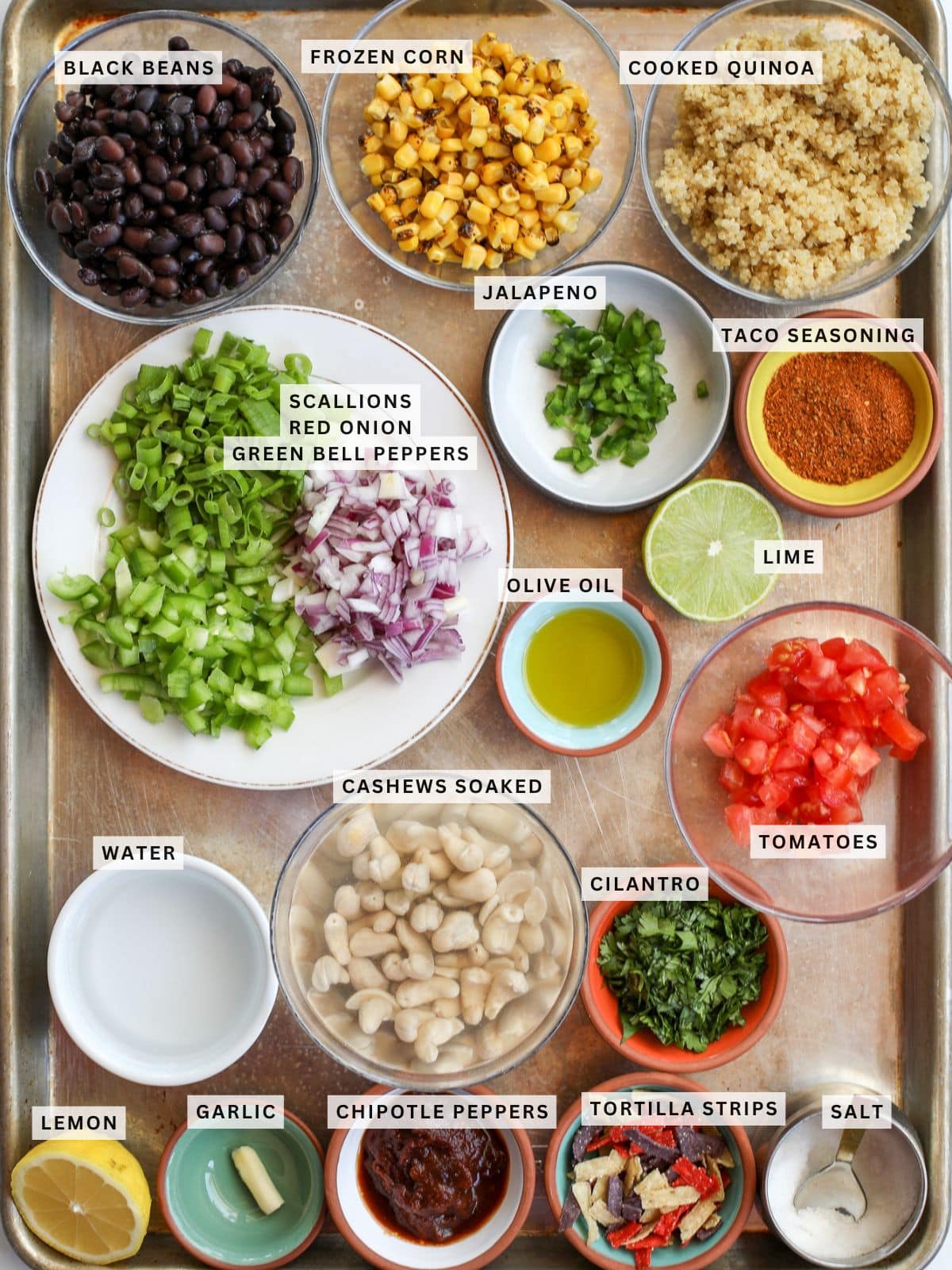 A tray with labeled bowls containing black beans, frozen corn, cooked quinoa, chopped vegetables, taco seasoning, olive oil, lime, soaked cashews, tomatoes, cilantro—everything needed for a delicious quinoa taco salad with chipotle aioli.