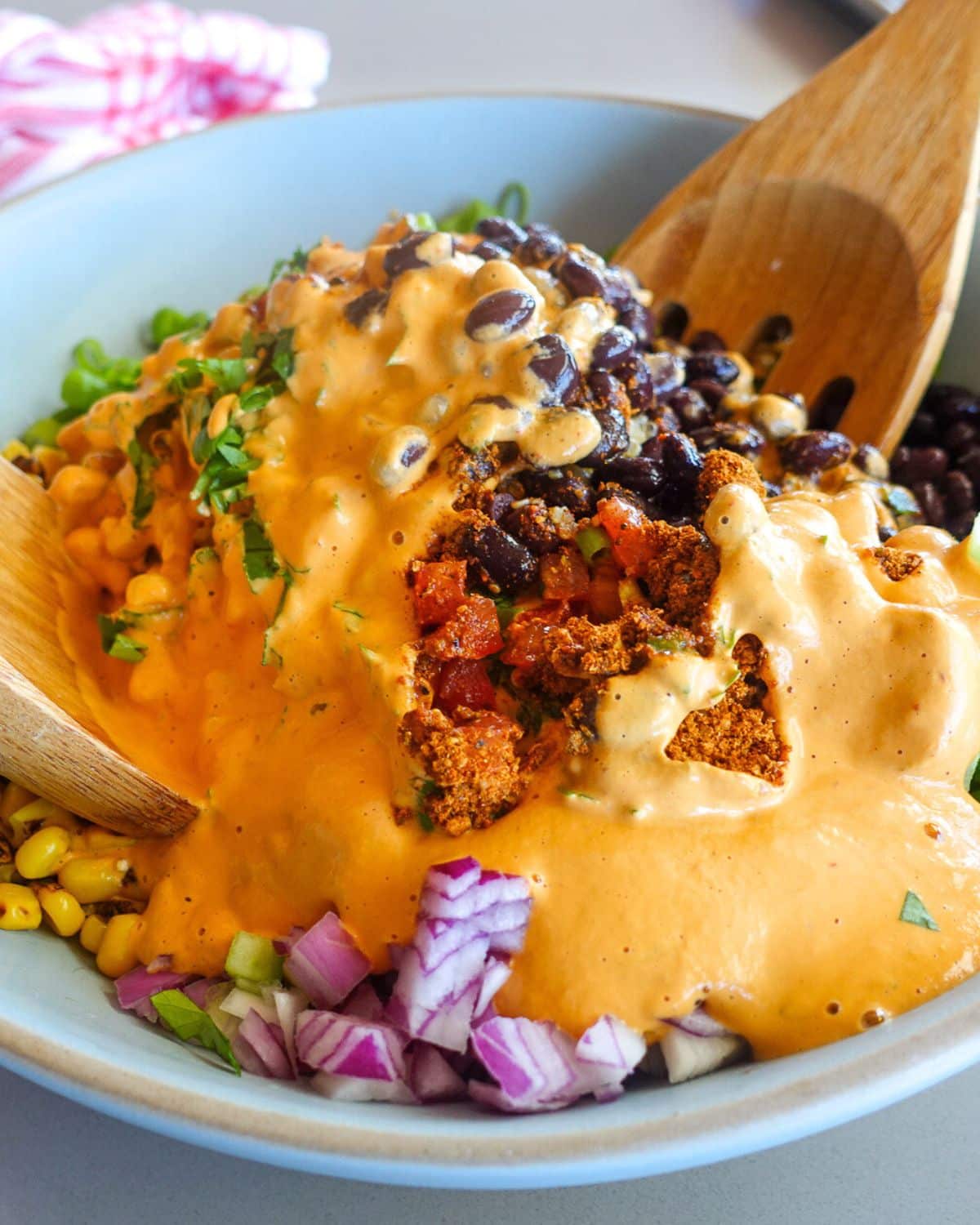 A bowl containing black beans, corn, chopped red onion, diced tomatoes, and creamy chipotle aioli is mixed with wooden spoons—perfect as a quinoa taco salad with bold flavors and vibrant colors.