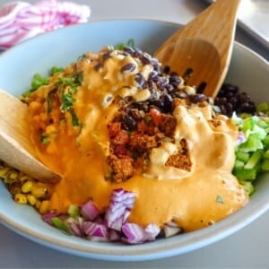 A bowl containing black beans, corn, chopped red onion, diced tomatoes, and creamy chipotle aioli is mixed with wooden spoons—perfect as a quinoa taco salad with bold flavors and vibrant colors.
