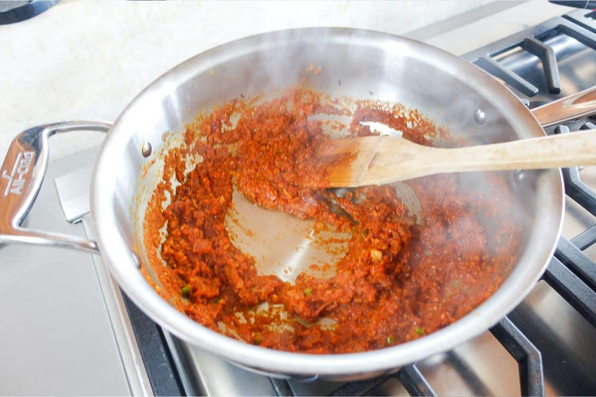 A stainless steel pan on a stove contains a thick red kadai paneer sauce being stirred with a wooden spoon, with steam rising from the mixture.
