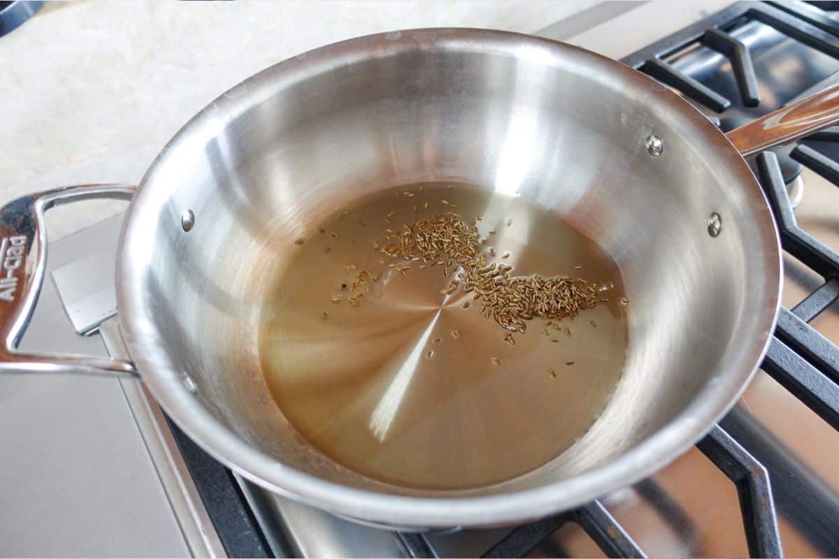 A stainless steel pan with oil and cumin seeds heating on a gas stove burner, ready for preparing delicious kadai paneer.