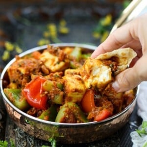 A hand dips roti into a bowl of kadai paneer in spicy masala, served in a metal dish with visible herbs and peppers.
