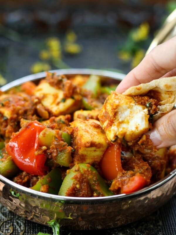 A hand dips roti into a bowl of kadai paneer in spicy masala, served in a metal dish with visible herbs and peppers.