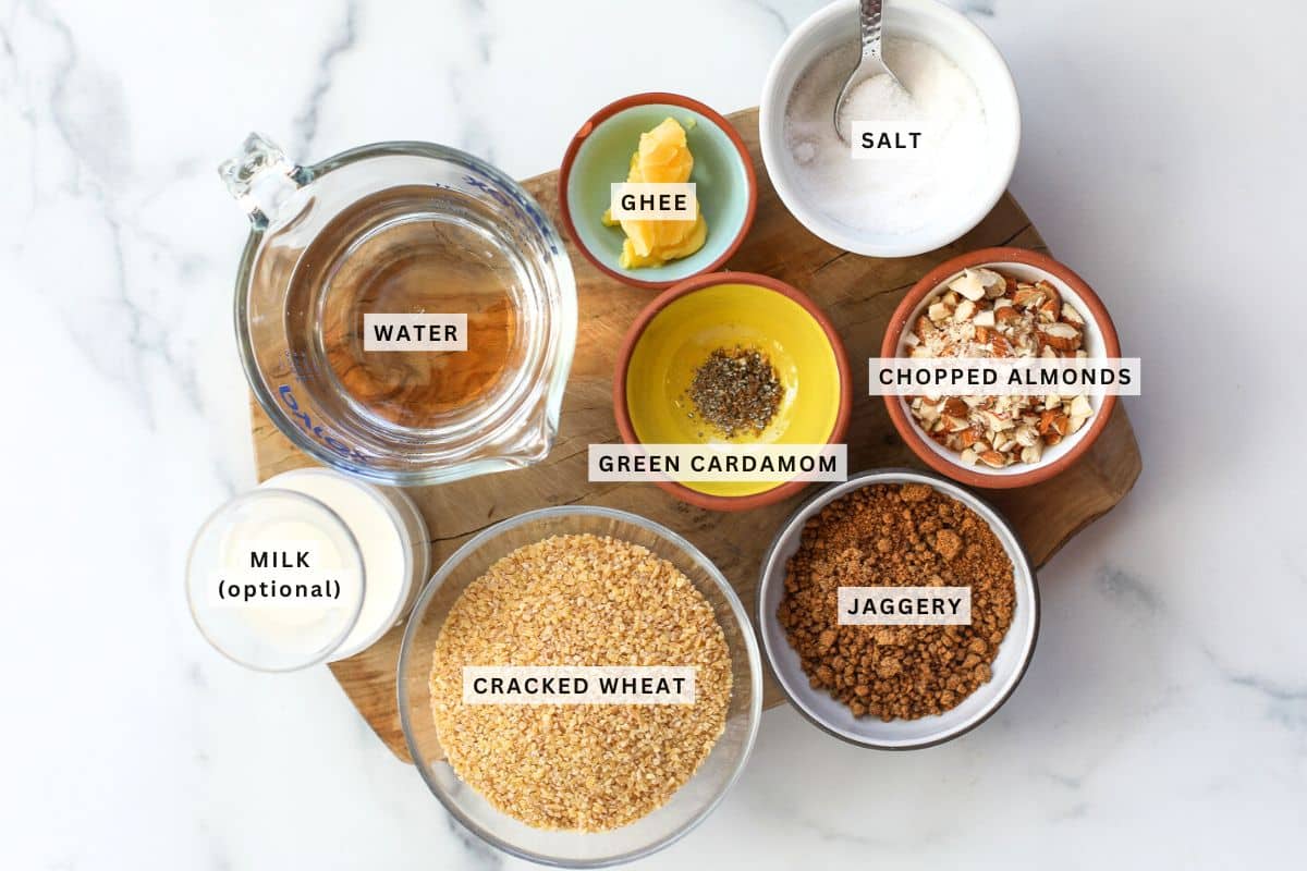 A top-down view of labeled ingredients for Sweet Daliya, including water, ghee, salt, chopped almonds, green cardamom, milk, cracked wheat, and jaggery on a wooden board.