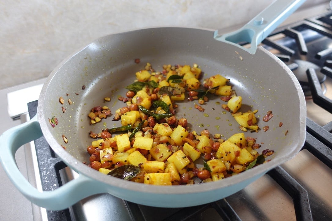Diced potatoes, peanuts, and spices sautéed in a blue non-stick pan on a gas stove—a classic touch for any aloo poha recipe.
