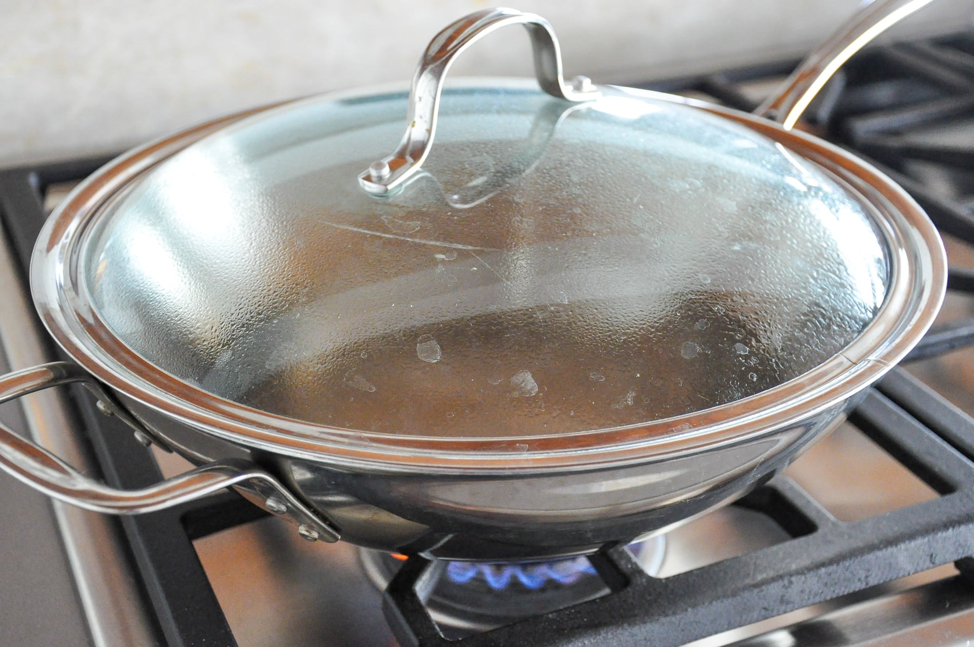A stainless steel pan with a glass lid sits on a gas stove burner with a blue flame steams inside and condensation collects on the lid.