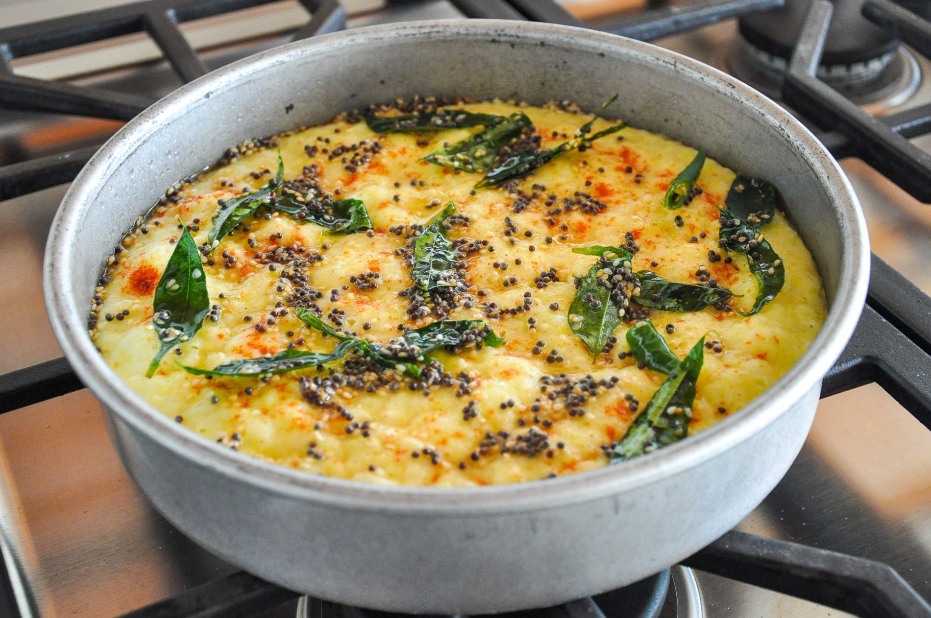 A round metal pan with cooked yellow rava dhokla, topped with green curry leaves and black mustard seeds, sits on a gas stove.