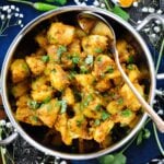 Jeera Aloo in a serving dish with a large spoon surrounded by fresh cilantro and spoonfuls of spices.